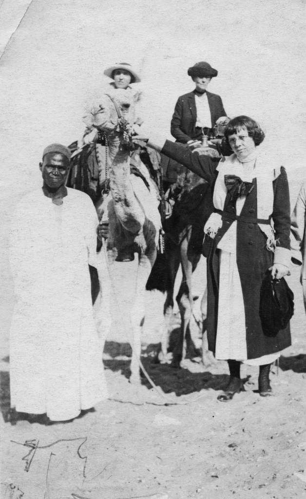 Two women sit on camels. Standing are an African man in traditional dress and a Euro-American woman.