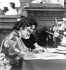 Louise Cross, sitting at a table in the capitol, signing the constitution. Next to her is a woman pointing at where to sign.