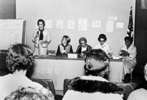 A woman speaks into a microphone. Four other panel members (all well-coifed women) sit at a head table facing the audience.