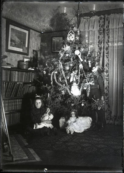 A girl hangs an ornament on a Christmas tree, and another girl poses in front of the tree holding a doll. On the left side of the photograph, four shelves of books can be seen and a painting above on the wall. Flooring is wood and two rugs are visible on the floor.