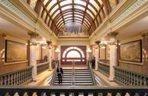 Picture of the grand staircase in the Montana State Capitol. The women's murals are on barely visible on either side of the staircase.