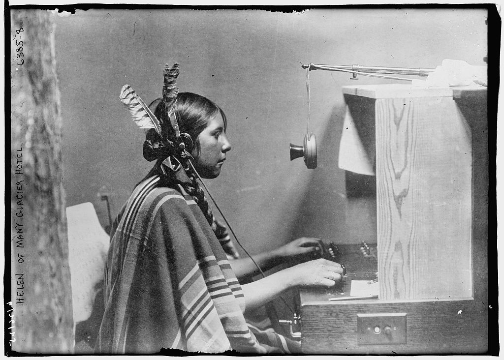 Blackfeet woman telephone operator, 1925, sitting at a hotel telephone exchange desk, wearing a blanket and feathers in her hair.