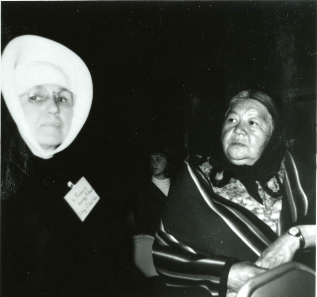 Sister Providencia, in full habit, with a nametag, sitting next to Florence Standing Rock, who is wearing a kerchief and blanket shawl