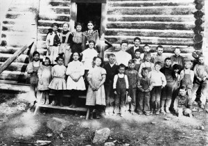School children (17 boys, most in overalls, and 9 girls) and teacher standing in front of a log school, 1907