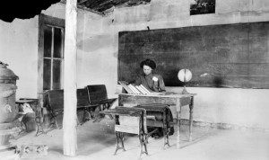 Teacher sits at a desk, with a globe and books inside a log school building. The interior also shows student desks, a wood stove, and a "blackboard," likely made of cloth.