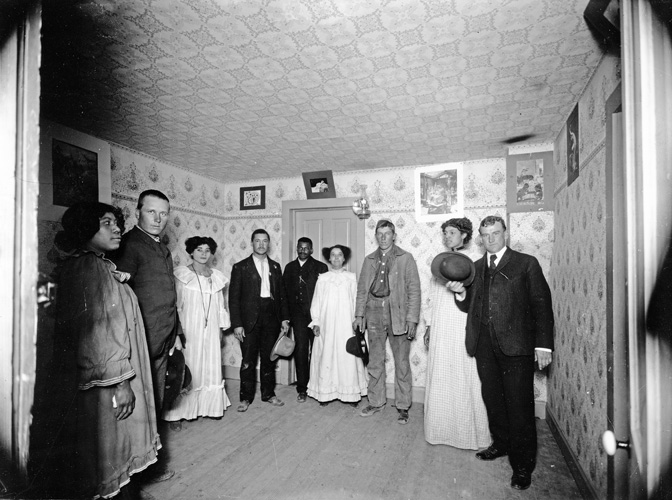 Four African American women in loosely fitting "Mother Hubbard" dresses pse with three white men and two African American men in a  wall-papered room with pictures on the wall.
