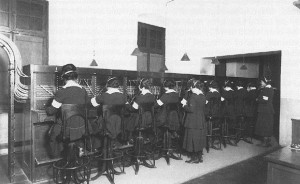 A row of women sitting at a switchboard. One woman supervisor is standing behind them. 