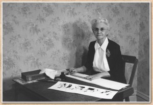 A gray haired woman sits at a table. An inked cartoon is in front of her.