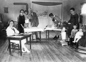 A makeshift doctor's office--probably in a school. A woman sits at a table (left.) Back left a female nurse holds a toddler while a male  doctor listens to his lungs with a stethoscope. Another woman holds a baby flat on a table, while a third woman  weights and measures a 3-4 year old on a doctor's scale. Two children sit in a rocking chair waiting.