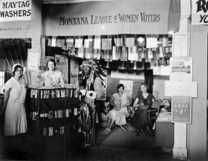 Montana League of Women Voters booth, decorated with flags of all nations . An Indian man in a ceremonial headdress stands at the counter, staffed by two young women. Two other women sit in the background.