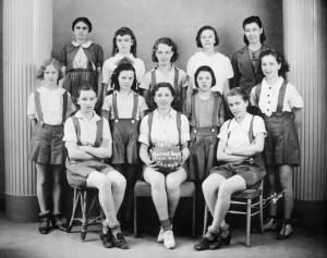 Team portrait of girls in uniform (overall shorts over white collared shirts) with their coaches. Girl sitting in center holds a ball labeled "1936. Sacred Heart Field Ball Champs"