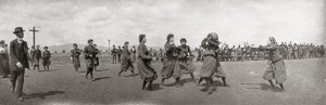 Women dressed in bloomers playing basketball on outside court. a man dressed in a suit (left) looks to be refereeing. Spectators sit on bleachers watching the game.