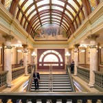 Picture of the grand staircase in the Montana State Capitol. The women's murals are on barely visible on either side of the staircase.